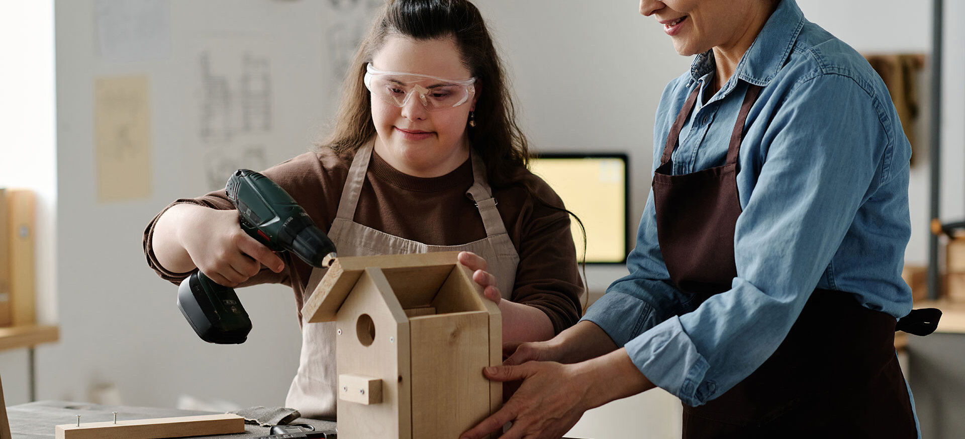 In einer Werkstatt baut eine junge Frau mit Schutzbrille und Akkuschrauber ein Holz-Vogelhaus zusammen, während eine Betreuerin daneben hilft.