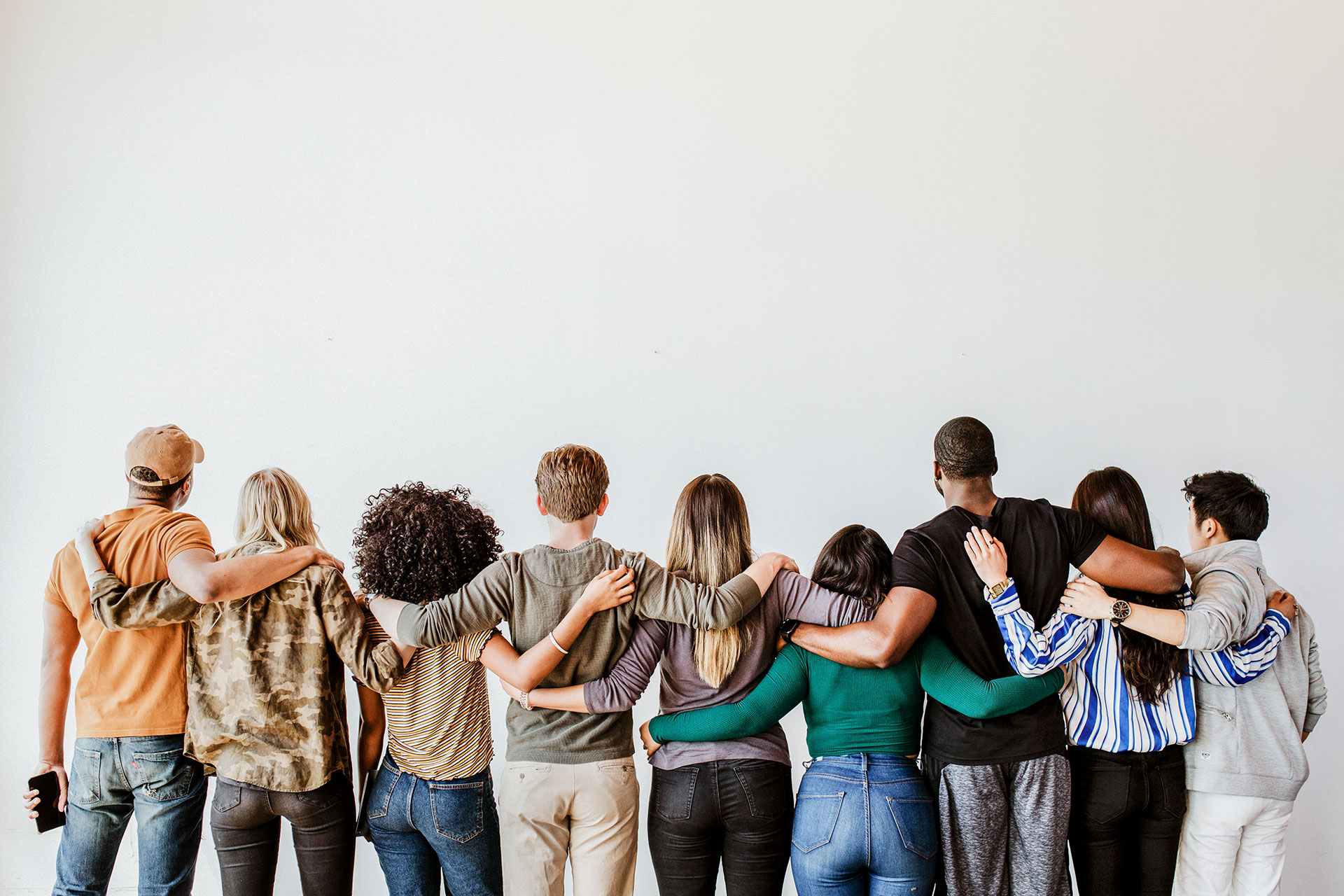 Eine Gruppe von neun jungen Menschen steht vor einer weißen Wand und legt sich gegenseitig die Arme um die Schultern.